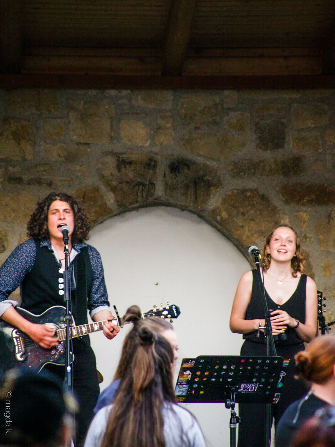 Auf dem Bild sind Charlotte und Tobi zu sehen. Tobi singt und spielt Gitarre. Charlotte lacht und hält etwas in der Hand.